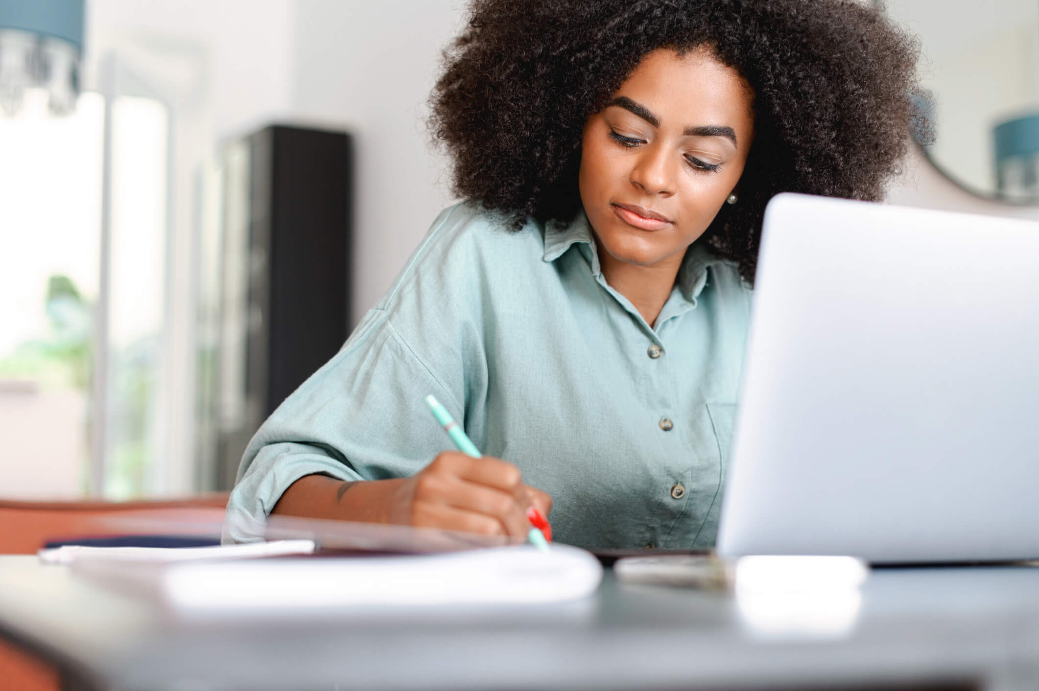 A woman is sitting at a desk in front of a laptop and is taking notes on paper; she has a content expression.