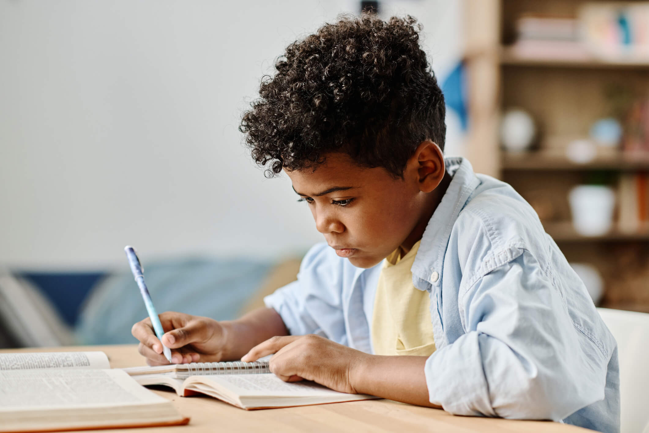 A boy is sitting at a desk and writing in a notebook; he is also reading a book with a focused expression.