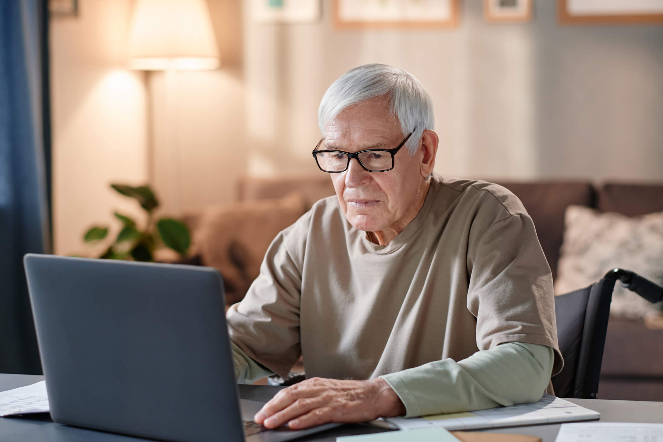 An older man in a wheelchair is in front of a laptop screen; he has a worried expression.