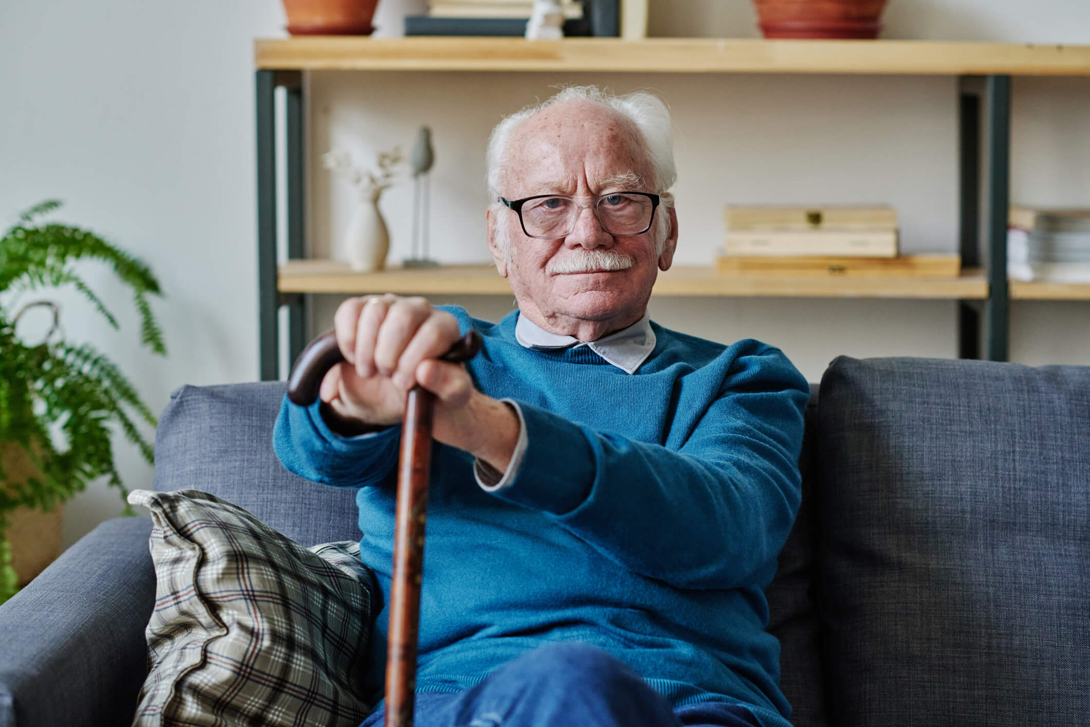 An older man is sitting on a couch and looking ahead; he is holding a cane and smiling.