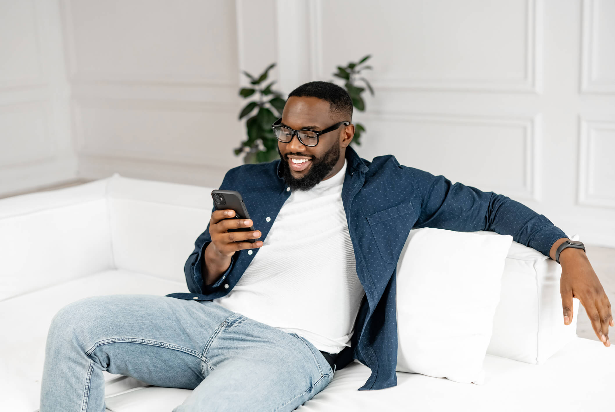 A bearded man with short dark hair, galsses, and a blue collared shirt over a white tee smiles at his phone as he sits on a white sofa.