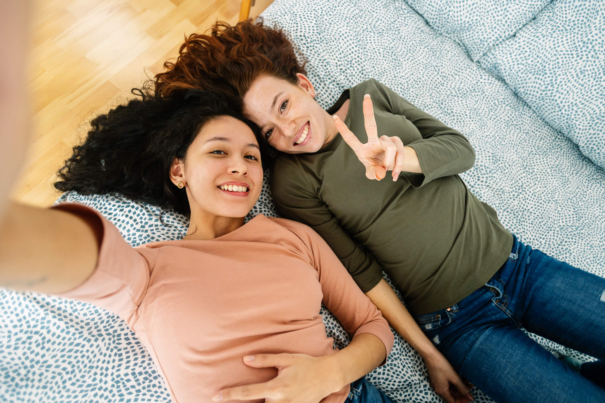 Two teens are lying on a bed, looking up at a camera, and taking a selfie; they are both smiling.