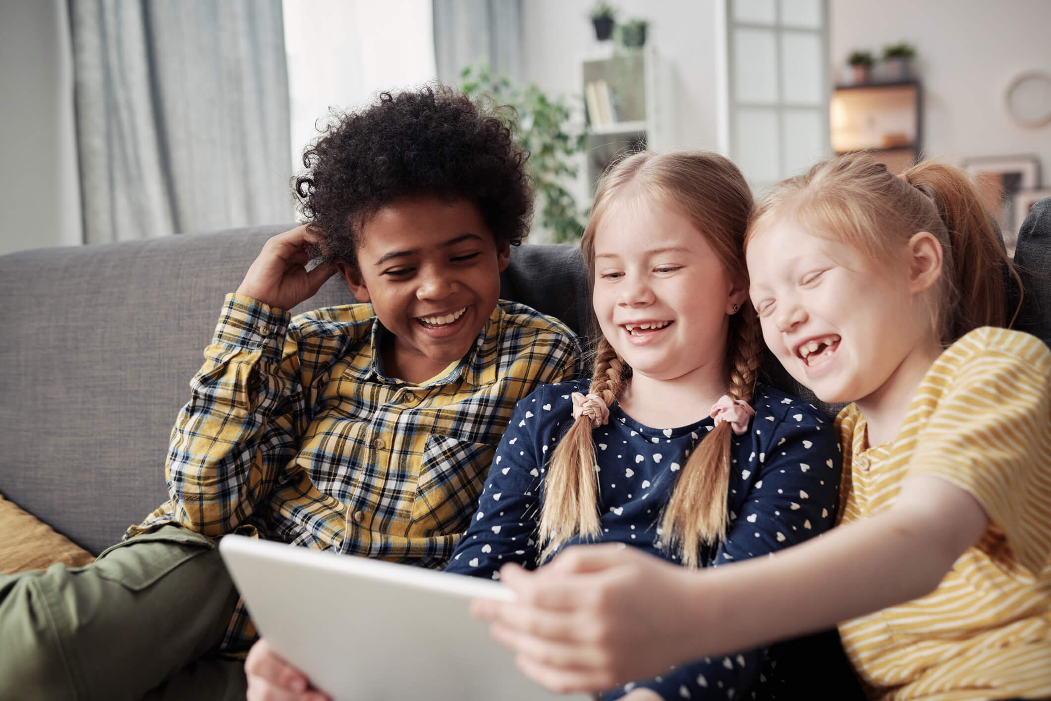 Three children are sitting on a couch together and looking at a tablet screen; they are all laughing and smiling.