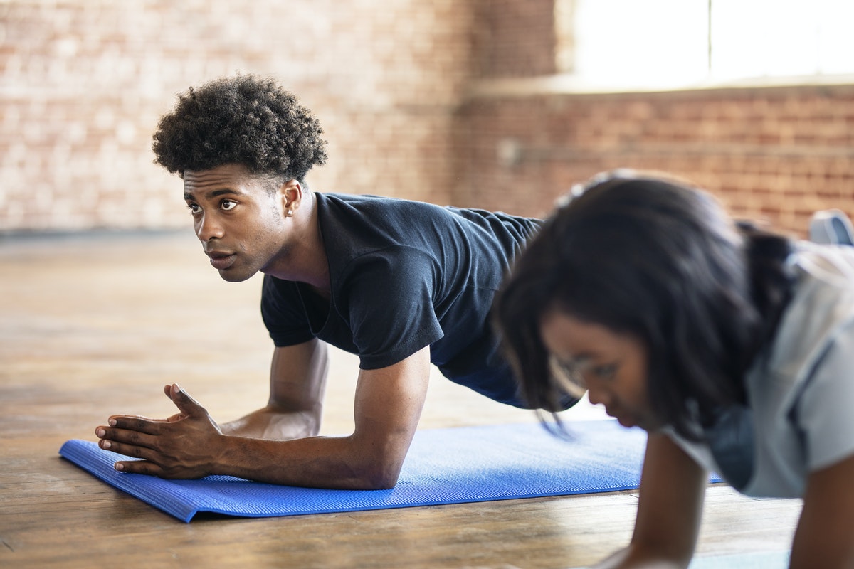 A man is in a yoga studio laying on a yoga mat next to another person; he has an amused expression.