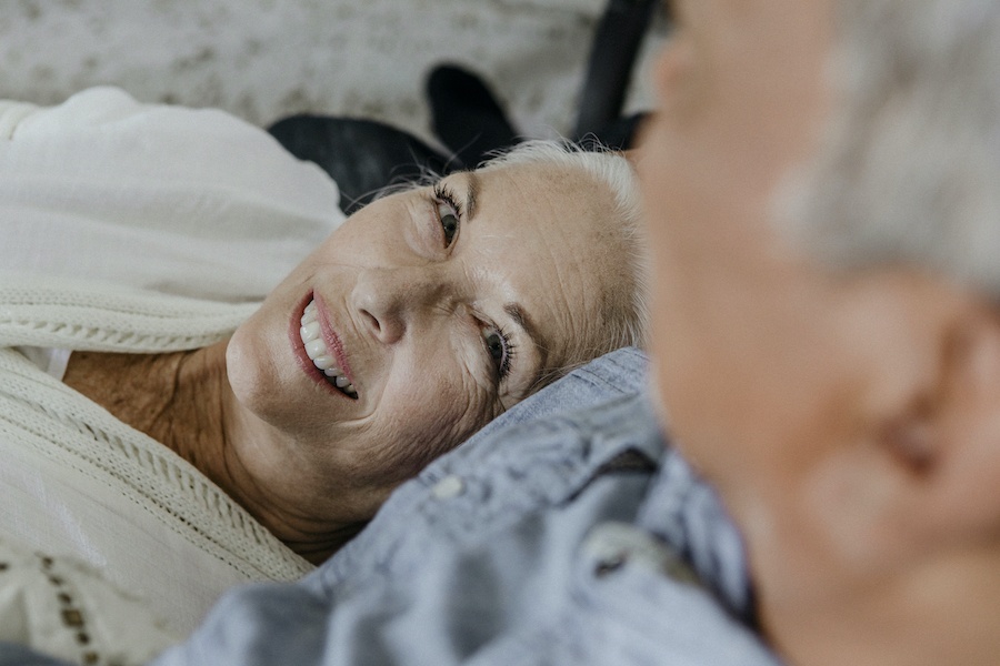 An elderly woman with white hair smiles as she looks up at her partner, an elderly man who is out of focus.